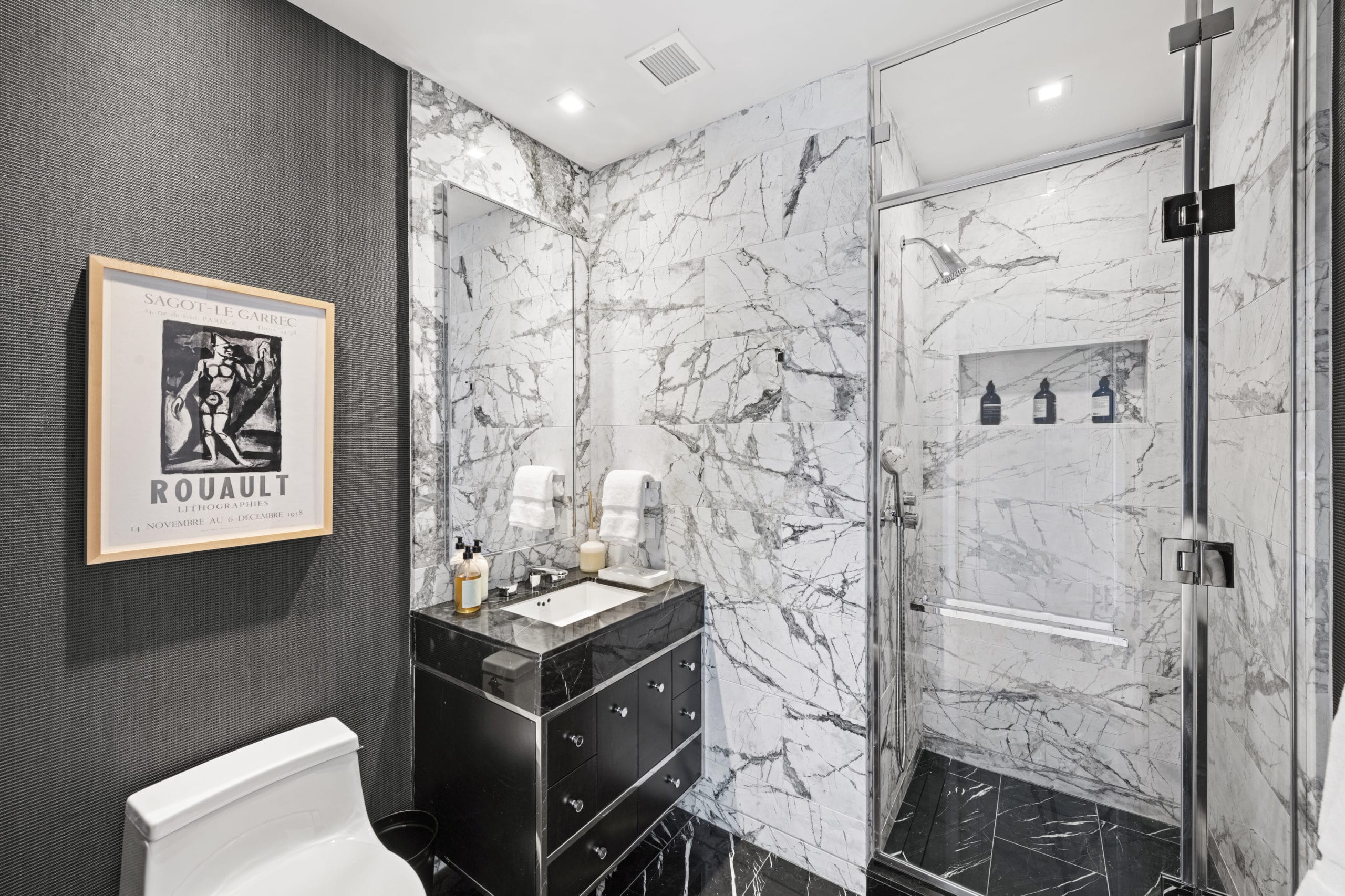 A condo bathroom with white marble shower, a vanity with mirror, and dark gray wall with a picture at The Centrale in NYC.