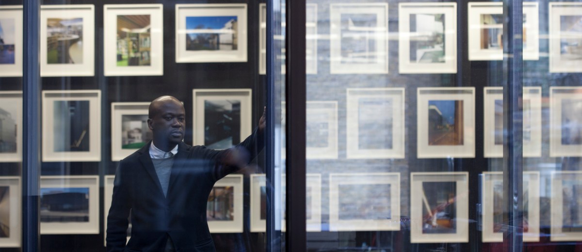 Portrait of David Adjaye looking out a window with his hand up on the wall. Framed pictures cover the walls behind him.