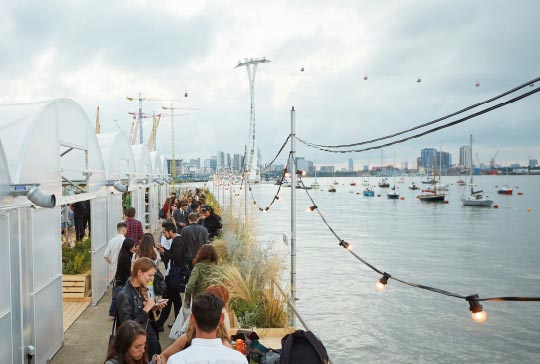 View of a crowded boardwalk with Thames river view in Greenwich Peninsula London. Has string lights and boats lined up.