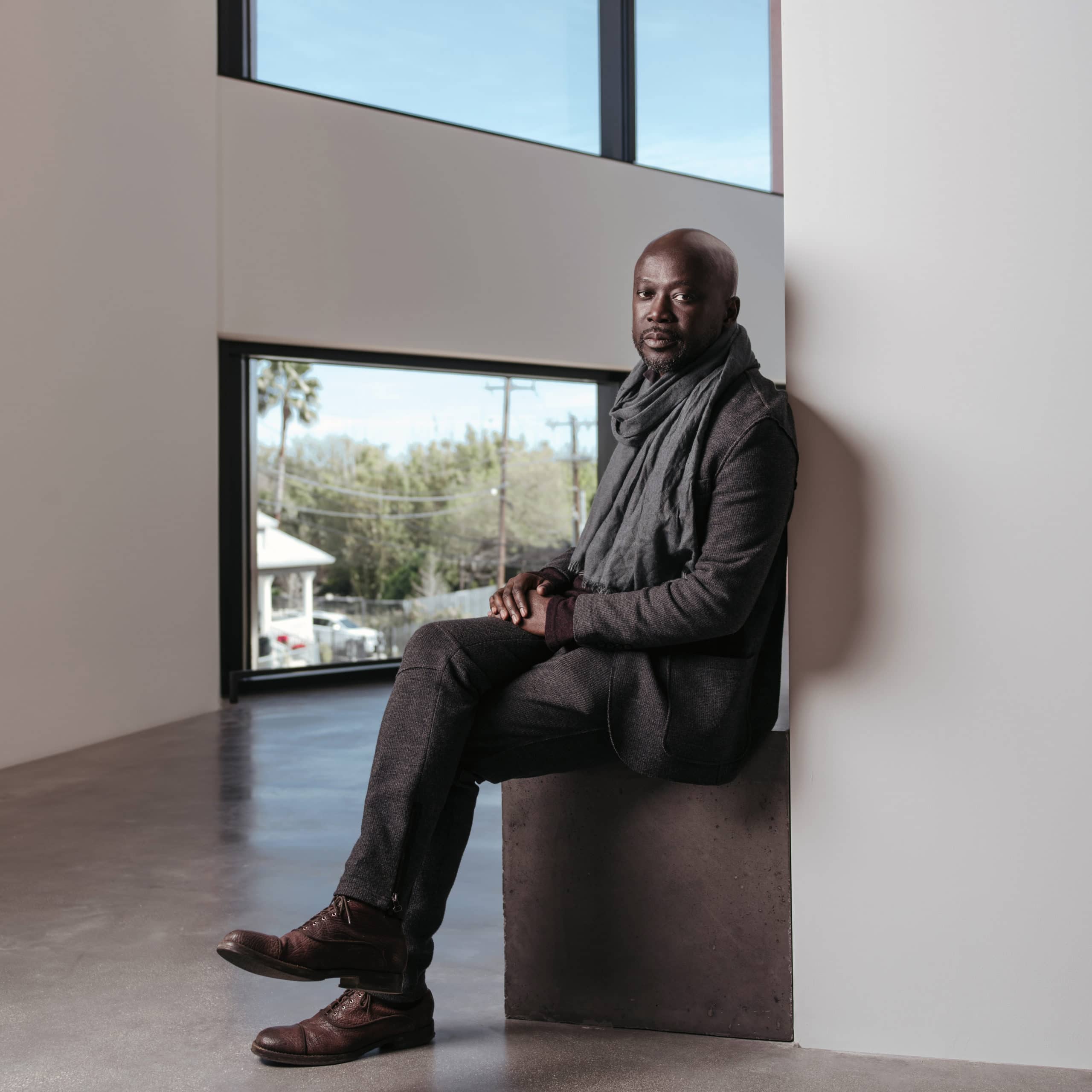 Profile picture of Sir David Adjaye by Josh Huskin. Man in jeans, jacket, and a scarf sitting with his legs crossed.