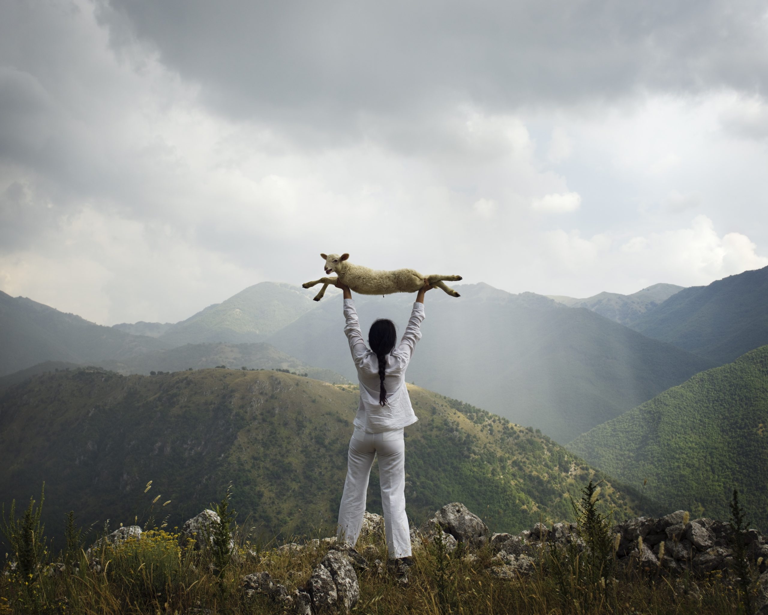 Picture of a woman holding a lamb above her head looking out at the mountains in the distance with dark clouds in the sky.