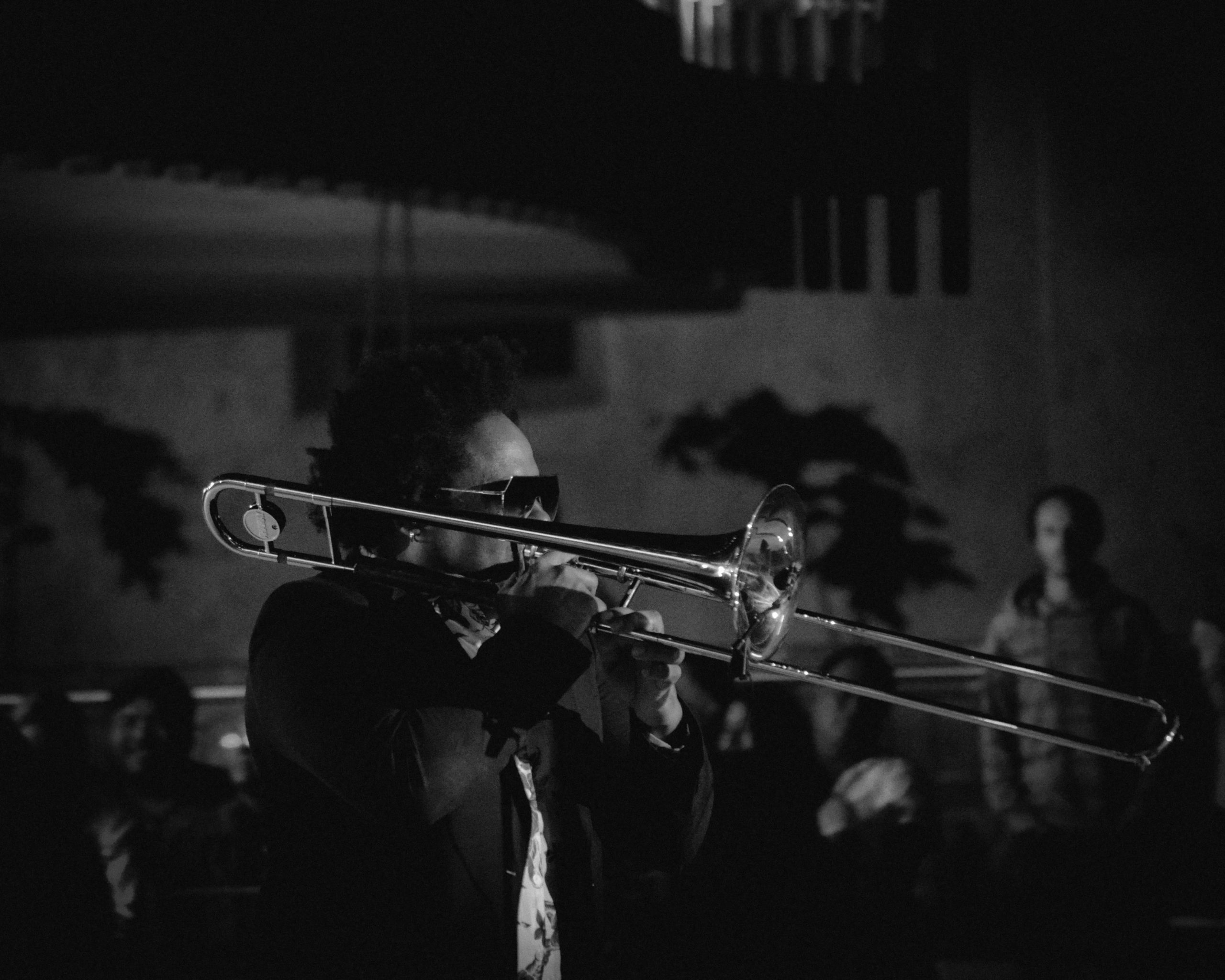 Black and white photo of musician playing the trombone. Side profile of the performer and audience members in the background.