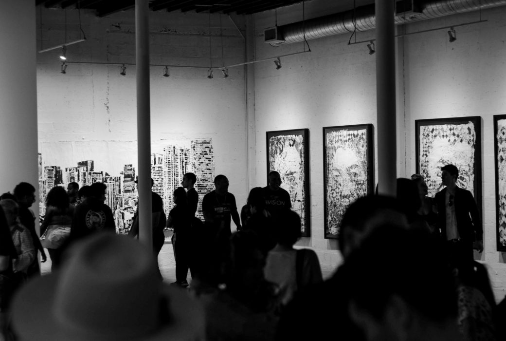 Black and white photo by Ruben Ramirez of a crowd at the Art Basel festival with three pictures and a mural on the far walls.
