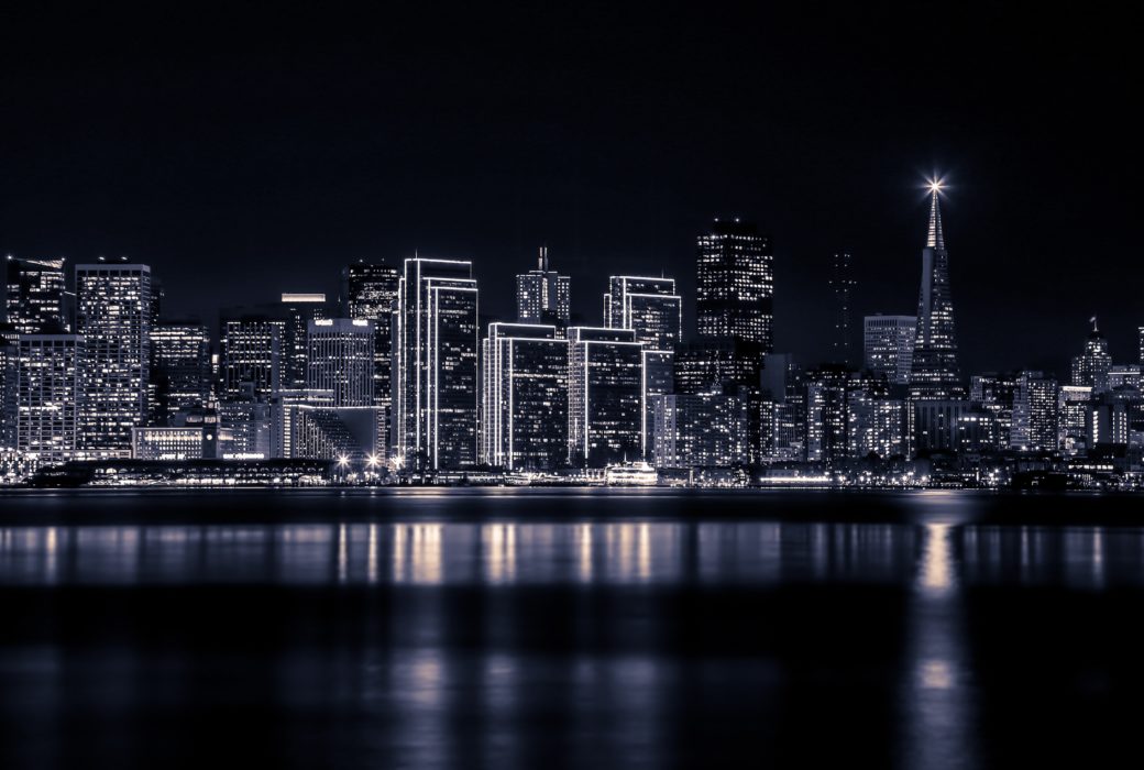 Black and white photo of San Francisco skyline at night. High rises on the bay with their images reflecting in the water.