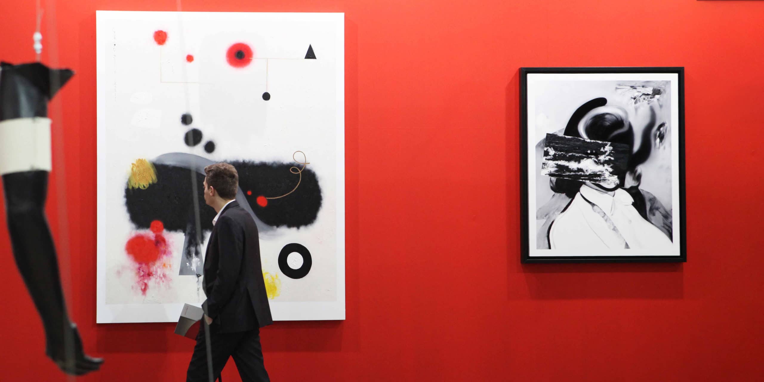Man walking past paintings hanging on a red wall. Sample exhibit from Italy's premier art fair, Artissima.