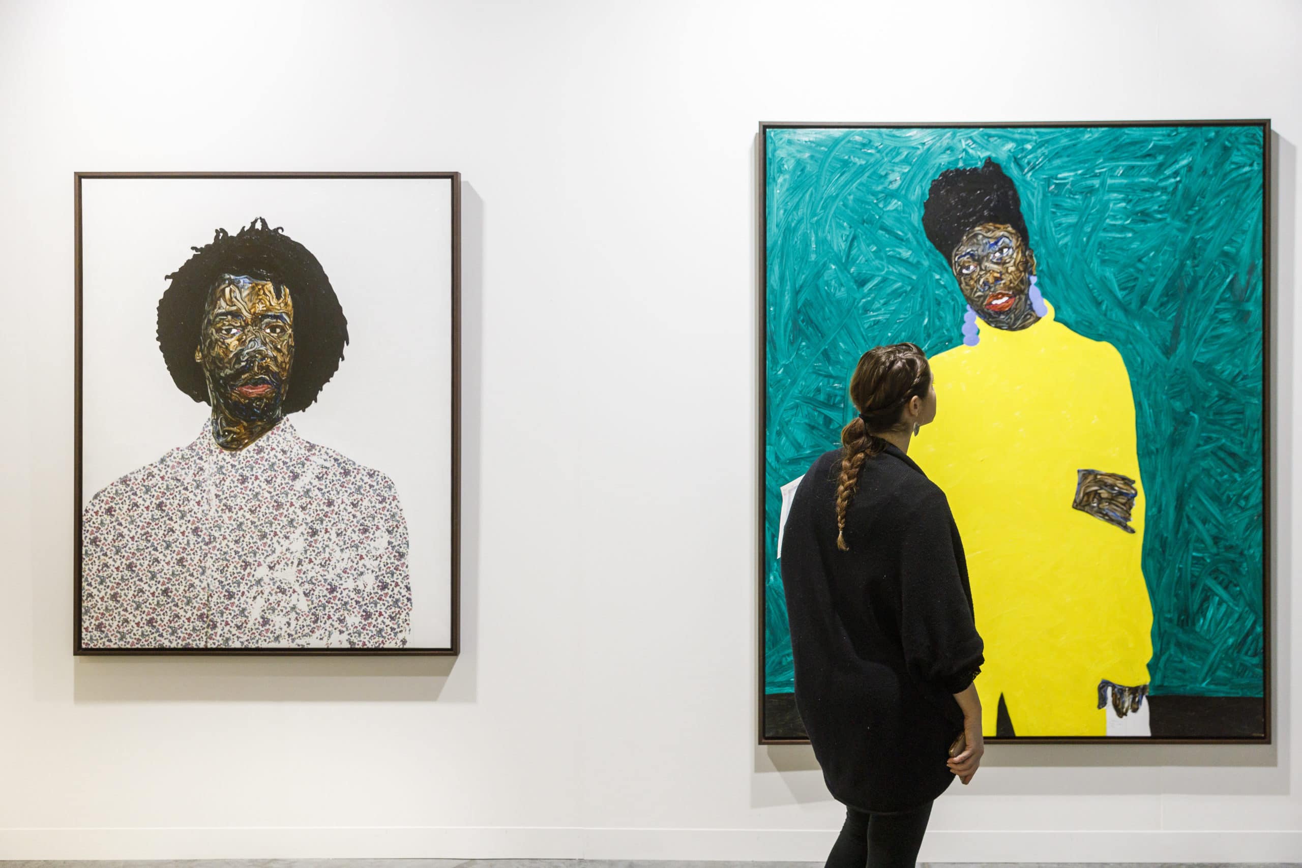 Closeup view of portrait paintings inside Art Basel. A woman is looking at a painting and has a white wall background.