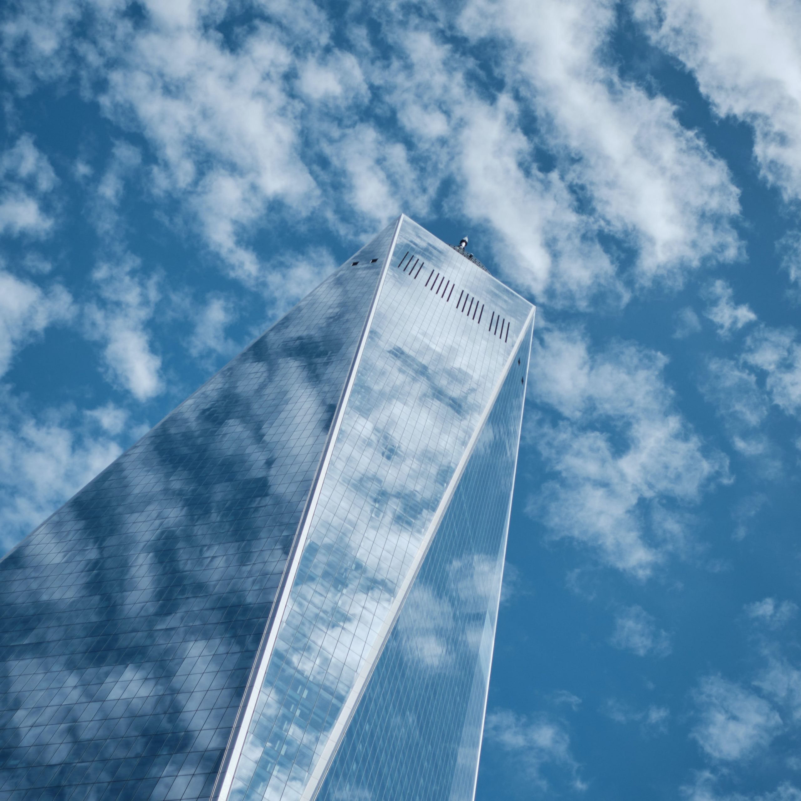 Birds eye view looking up at a glass skyscraper and clouds. Blue sky, clouds, and sun are reflecting off of the glass walls.