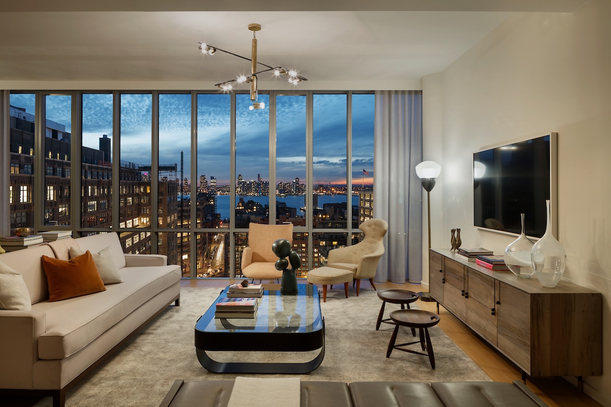 Interior view of 565 Broome residence living room with window view of New York City. Has full furniture, white walls and TV.